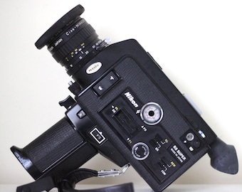 May include: A black Nikon R8 Super 8mm movie camera featuring a Cine-Nikkor lens, a textured grip, and various control switches. The camera body displays the Nikon logo, a PASSED sticker, and the text R8 SUPER MADE IN JAPAN on the side panel.