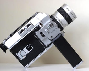 Può includere: Una cinepresa Super 8 color argento e nero con obiettivo zoom. Il dispositivo è dotato di un'impugnatura laterale, un interruttore manuale e automatico, una finestra per l'indicazione del tipo di pellicola e un mirino. L'obiettivo reca la scritta Canon Zoom Lens.