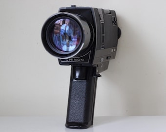 May include: A black Chinon Super 8 movie camera featuring a prominent circular lens with a reflective glass surface, a textured pistol-grip handle, and a rectangular body design. The brand name Chinon is printed in white lettering on the front.