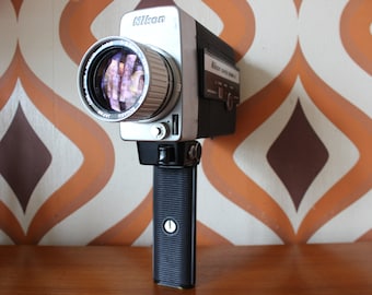 May include: A vintage Nikon Super 8 movie camera with a silver body and black handle. The lens is prominent, reflecting the surroundings. The camera has the Nikon logo and other text. The handle has a textured grip and a button.