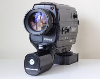 May include: A black Bauer movie camera featuring a prominent front lens with a reflective glass surface, a side-mounted handle with the text Robert Bosch GMBH, and various control dials and buttons on the main body, including the Bauer brand name.