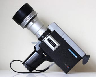 May include: Vintage Canon Super 8 movie camera with a black body, silver lens, and black handle. The camera has a black lens cap and a silver lens with measurement markings. A black strap is attached to the handle.