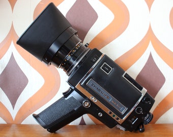 May include: A vintage black Chinon 812 Macro movie camera with a large lens and handle. The camera has silver trim and is angled towards the viewer. The background features a retro orange and brown pattern.