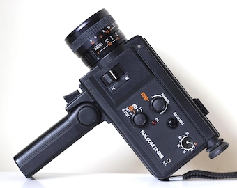May include: Black Nalcom EX-5000 vintage movie camera with a lens and handle. The camera has various dials, switches, and the text "NALCOM EX-5000 R.C." on the front. A black strap is attached to the bottom.