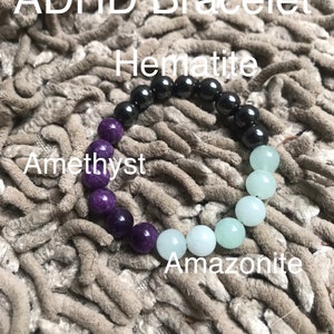 May include: A bracelet made of amethyst, amazonite, and hematite beads. The bracelet is on a fuzzy, gray surface. The text "ADHD Bracelet" is at the top of the image. The text "Hematite" is above the hematite beads. The text "Amethyst" is below the amethyst beads. The text "Amazonite" is below the amazonite beads.