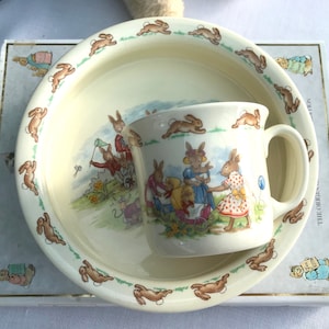 May include: A white ceramic children's bowl and mug with a Peter Rabbit design. The bowl has a green rim and a border of brown rabbits. The mug has a handle and a picture of Peter Rabbit and his friends.