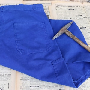Può includere: Pantaloni da lavoro blu brillante con ampie tasche e un piccone. Il piccone ha un manico in legno e una testa in metallo. I pantaloni sono in tessuto resistente. Gli oggetti sono su un vecchio giornale.