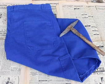 May include: Bright blue work pants with large pockets and a pickaxe resting on top. The pants are made of a durable fabric, and the pickaxe has a wooden handle and a metal head. The items are placed on top of old newspaper.