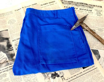 May include: Bright blue work pants with a large pocket, laid on top of vintage French newspapers. A pickaxe rests on the pants. The newspapers have black and white images and text.