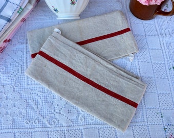 May include: Two linen tea towels with a single red stripe down the center. The towels are folded and laid on a white crocheted tablecloth.