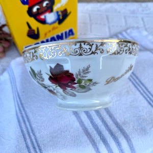 Puede incluir: Un tazón de porcelana blanca con un borde dorado y un diseño floral. El tazón tiene una inscripción dorada que dice "Danville".