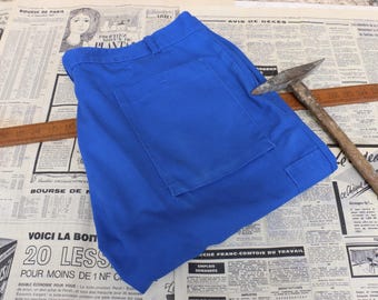 May include: Bright blue work pants with a large back pocket and a smaller side pocket. A vintage pickaxe with a wooden handle rests on the pants. The background is a vintage newspaper.