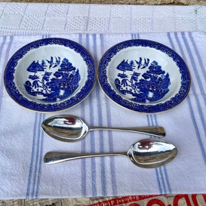 Pode incluir: Duas tigelas de porcelana azul e branca com borda dourada e um design de salgueiro. Duas colheres de prata são colocadas na frente das tigelas.