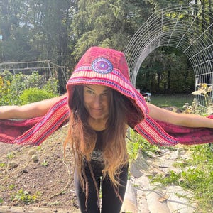 May include: A hooded scarf in shades of red, pink, and blue. The hood features a circular design with a blue and purple center. The scarf extends into long, fringed sleeves. The person is wearing a black top and black leggings.