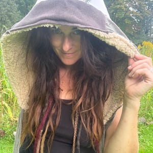 May include: A hooded garment featuring a cream-colored hood with a brown mushroom and pine tree print, a dark brown waffle-knit trim, and a thick, beige sherpa fleece lining. The garment is worn over a simple black tank top, showcasing the textures.
