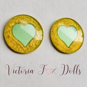 Puede incluir: Dos cabujones redondos de resina amarilla con un dise&ntilde;o de coraz&oacute;n verde claro. Los cabujones tienen un di&aacute;metro de aproximadamente 2,5 cm.