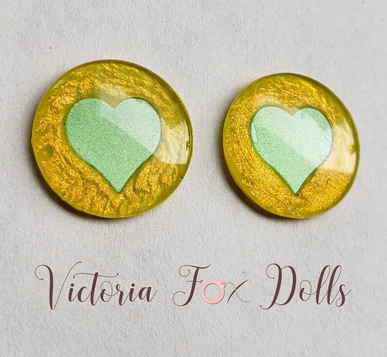 Puede incluir: Dos cabujones redondos de resina amarilla con un dise&ntilde;o de coraz&oacute;n verde claro. Los cabujones tienen un di&aacute;metro de aproximadamente 2,5 cm.