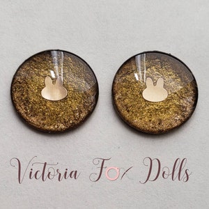 May include: Two round, gold-colored resin doll eyes with a white bunny silhouette in the center. The eyes have a glittery finish.