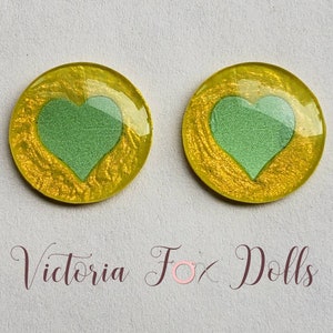 Puede incluir: Dos cabujones redondos de resina amarilla con un dise&ntilde;o de coraz&oacute;n verde. Los corazones est&aacute;n ligeramente elevados y tienen una superficie texturizada. Los cabujones tienen un di&aacute;metro de aproximadamente 2,5 cm.