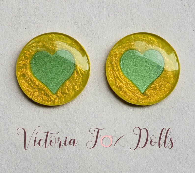 Puede incluir: Dos cabujones redondos de resina amarilla con un dise&ntilde;o de coraz&oacute;n verde. Los corazones est&aacute;n ligeramente elevados y tienen una superficie texturizada. Los cabujones tienen un di&aacute;metro de aproximadamente 2,5 cm.