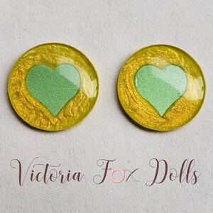 Puede incluir: Dos cabujones redondos de resina amarilla con un dise&ntilde;o de coraz&oacute;n verde. Los corazones est&aacute;n ligeramente elevados y tienen un acabado brillante.
