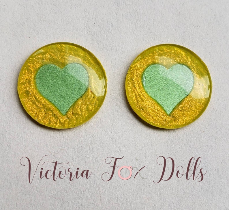 Puede incluir: Dos cabujones redondos de resina amarilla con un dise&ntilde;o de coraz&oacute;n verde. Los corazones est&aacute;n ligeramente elevados y tienen un acabado brillante.