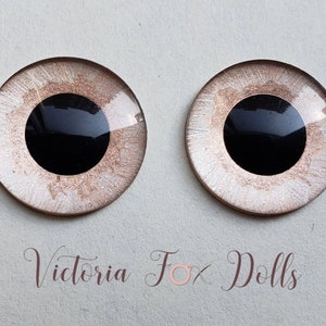 Puede incluir: Dos ojos de muñeca redondos de vidrio con una pupila negra y un iris blanco con un brillo marrón. Los ojos miden aproximadamente 2,5 cm de diámetro.