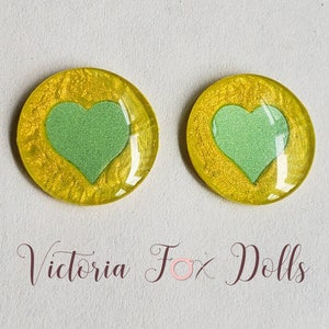 Puede incluir: Dos cabujones redondos de resina amarilla con un dise&ntilde;o de coraz&oacute;n verde. Los corazones tienen un acabado brillante.