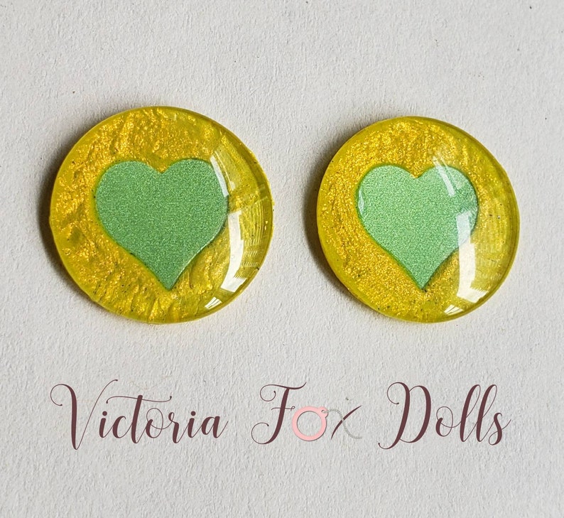 Puede incluir: Dos cabujones redondos de resina amarilla con un dise&ntilde;o de coraz&oacute;n verde. Los corazones tienen un acabado brillante.