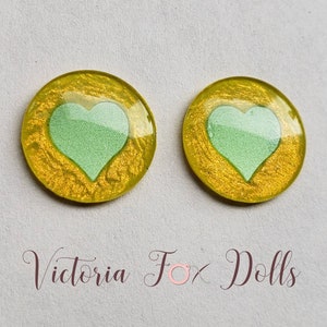 Puede incluir: Dos cabujones redondos de resina amarilla con un dise&ntilde;o de coraz&oacute;n verde. Los cabujones tienen un acabado brillante.