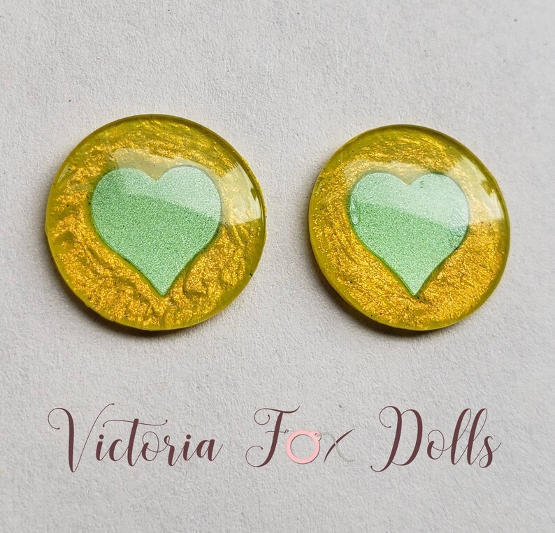 Puede incluir: Dos cabujones redondos de resina amarilla con un dise&ntilde;o de coraz&oacute;n verde. Los cabujones tienen un acabado brillante.