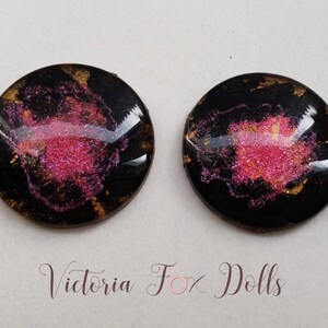 Peut inclure: Deux cabochons ronds en r&eacute;sine noire avec des paillettes roses et dor&eacute;es incrust&eacute;es dans la r&eacute;sine. Les cabochons mesurent environ 2,5 cm de diam&egrave;tre.