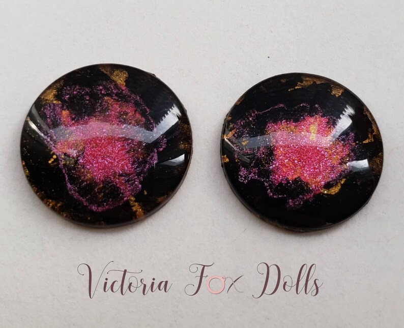 Peut inclure: Deux cabochons ronds en r&eacute;sine noire avec des paillettes roses et dor&eacute;es incrust&eacute;es dans la r&eacute;sine. Les cabochons mesurent environ 2,5 cm de diam&egrave;tre.