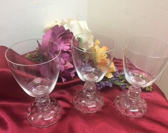 May include: Three clear glass wine glasses with a textured base. The glasses are arranged on a red surface with a bouquet of pink, white, and yellow flowers.