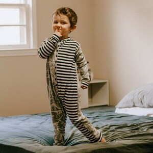 Può includere: Un bambino con un pigiama intero a righe grigie e nere. Il pigiama ha un motivo di dinosauri su un lato e strisce bianche e nere sull'altro. Il bambino è in piedi su un letto con una trapunta blu.