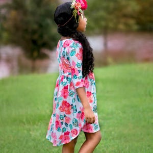 May include: A young girl wearing a floral print dress with a pink and green color scheme. She is wearing a flower crown and brown shoes. The girl is standing in a grassy field.