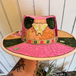 May include: A pink, wide-brimmed hat with a patchwork design. The hat features a butterfly applique, floral trim, and a patterned pink fabric. The brim is a light beige color, and the hat has a decorative gold trim.