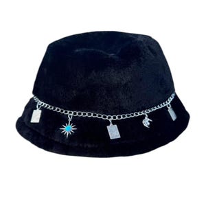 May include: Black fuzzy bucket hat with a silver chain and charms. The charms include a star with a turquoise stone, a square, and a crescent moon.