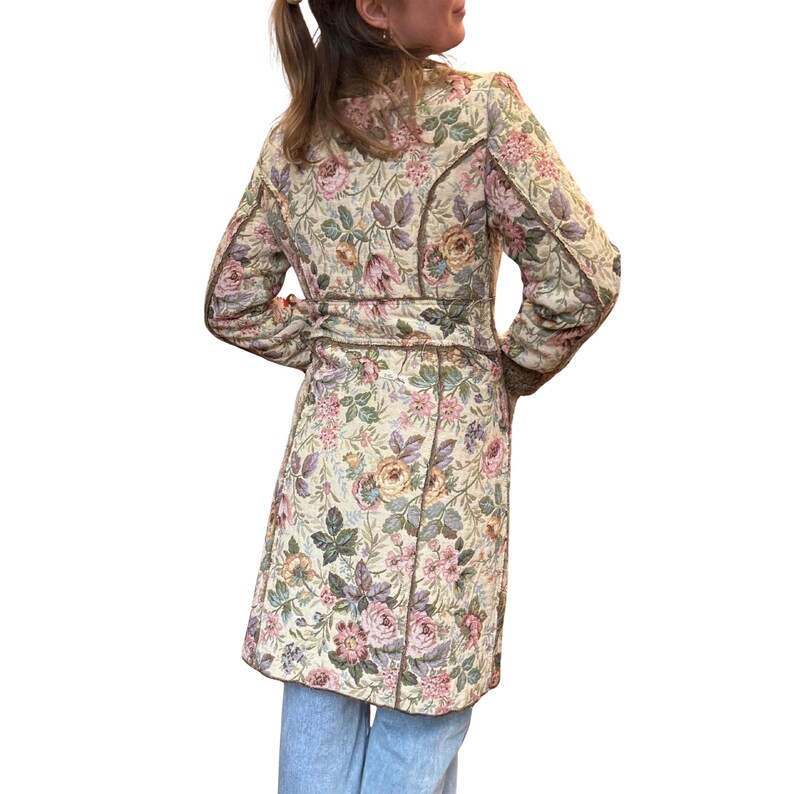 May include: A long, floral-patterned coat in cream, pink, and green. The coat features a belted waist and brown trim. The floral design includes roses and other flowers. The coat is made of a quilted fabric.