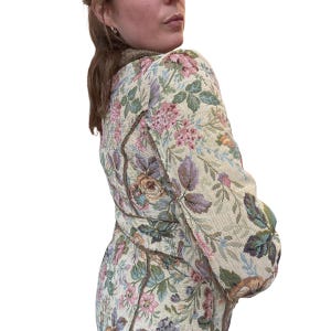 May include: A floral tapestry jacket with a cream background and a pattern of pink, purple, and green flowers and leaves. The jacket has a brown trim and a high collar.