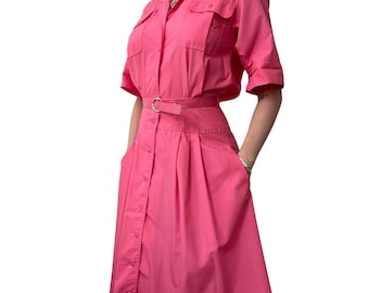 May include: A bright pink dress with short sleeves, a collared neckline, and a button-down front. The dress features two chest pockets, a matching belt with a silver buckle, and a pleated skirt. The dress has a classic, vintage style.
