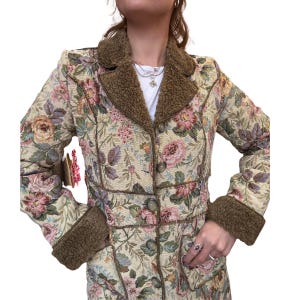 May include: A floral tapestry coat with a brown faux fur collar and cuffs. The coat features a button-down front and a detailed floral pattern in shades of pink, green, and beige. The coat has a vintage aesthetic.