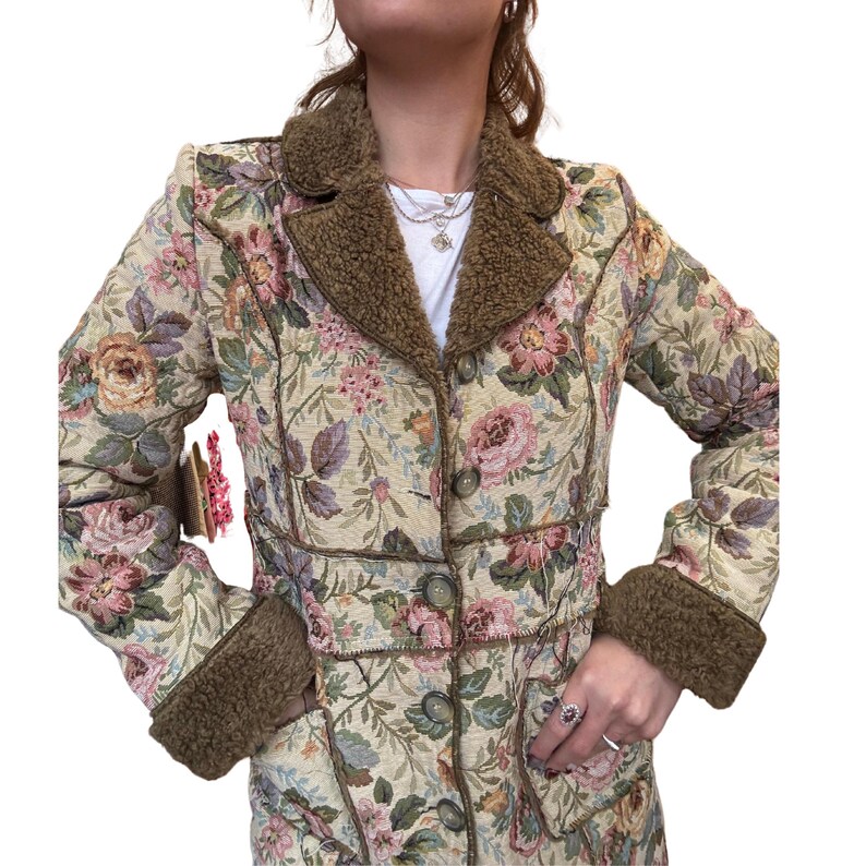 May include: A floral tapestry coat with a brown faux fur collar and cuffs. The coat features a button-down front and a detailed floral pattern in shades of pink, green, and beige. The coat has a vintage aesthetic.