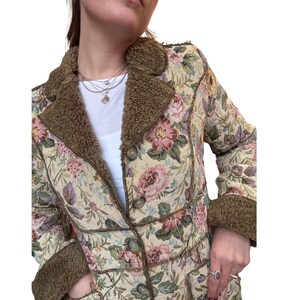 May include: A floral tapestry coat with a brown faux fur collar and trim. The coat features a cream background with pink and green floral patterns, and is buttoned up. The coat has a vintage aesthetic.
