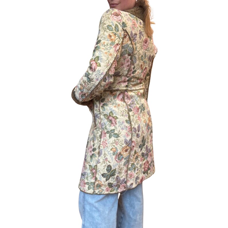 May include: A floral print coat in a light beige color with pink, purple, and green floral patterns. The coat has long sleeves and a belt at the waist. The coat is knee-length and has a classic design.