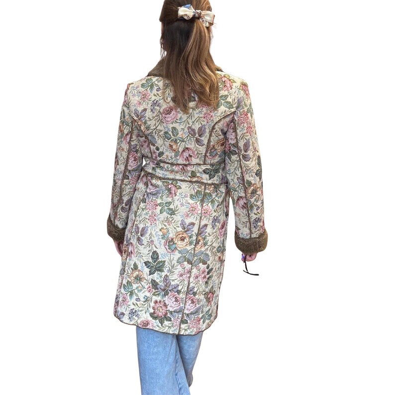 May include: A floral print coat with a belt and brown trim. The coat features a cream-colored background with pink, purple, and green floral patterns. The coat has a brown fur collar and cuffs. The person is wearing blue jeans.