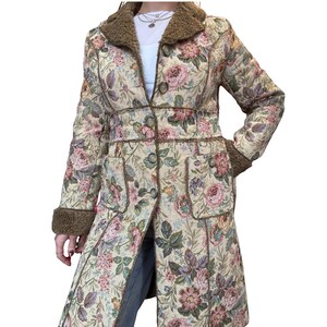 May include: A long, floral-patterned coat with a brown faux fur collar and cuffs. The coat features a cream-colored background with pink, purple, and green floral designs. It has a button-up front and two front pockets.