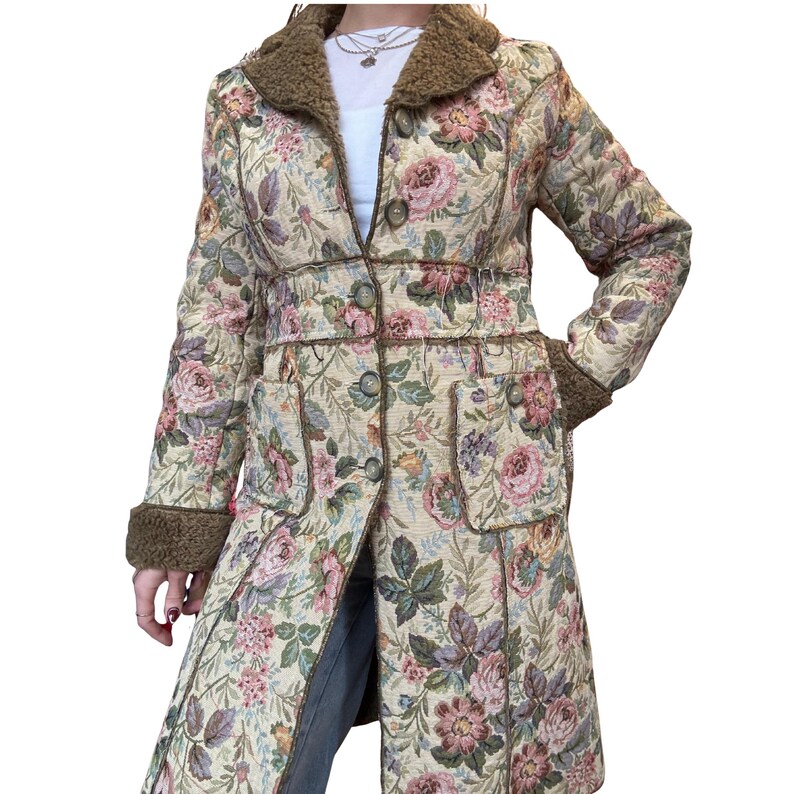 May include: A long, floral-patterned coat with a brown faux fur collar and cuffs. The coat features a cream-colored background with pink, purple, and green floral designs. It has a button-up front and two front pockets.