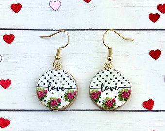 Può includere: Un paio di orecchini pendenti dorati con sfondo bianco e nero a pois e un design floreale con la parola "love" in lettere nere.