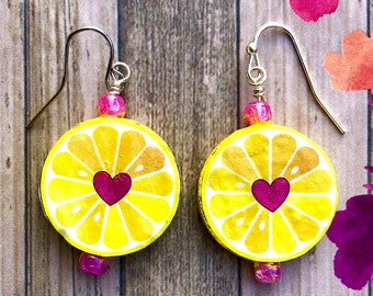 Può includere: Un paio di orecchini pendenti con ciondoli circolari a forma di fetta di limone gialla, caratterizzati da un cuore rosa scuro al centro. Ogni orecchino è decorato con una piccola perla rosa traslucida sopra e sotto la fetta di limone.